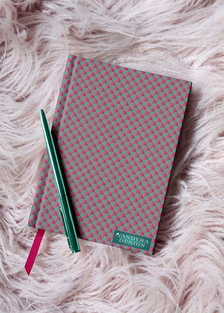 Notizbuch liegt auf Fellteppich. Muster ist ein neonfarbenes Schuppenmuster in Grün und Pink. Dekoelement ein grüner Metallkugelschreiber. Auf dem Bild ist das Label - der Schriftzug Vandyra Design von der Designerin zusehen.
